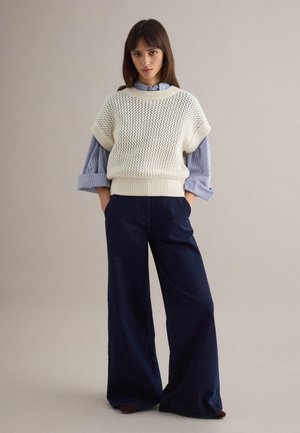 Mujer joven de pie con las manos en los bolsillos, vistiendo un chaleco de punto blanco sobre una camisa de rayas azules y pantalones de pierna ancha azul oscuro.
