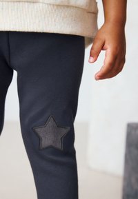 Leggings gris oscuro con un aplique texturizado en forma de estrella en la rodilla izquierda. La tela parece suave y elástica, complementando una camiseta clara con motas.