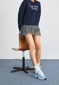 Felpa blu navy con il testo "Pepe Jeans London", abbinata a una mini gonna floreale con volant, calzini bianchi e sneakers New Balance azzurro chiaro.