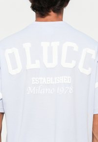 Person med en lyseblå skjorte med store hvite bokstaver "OLUCC" og mindre tekst "ETABLERET Milano 1978" på ryggen.