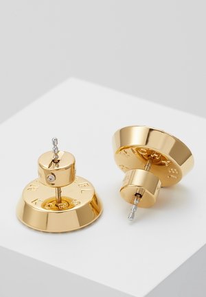 Paar glänzende goldene Ohrstecker mit runden Grundflächen, eingraviertem Markennamen und kleinen klaren Edelstein-Akzenten auf weißer Oberfläche.