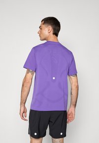 Lila Sport-T-Shirt, kurzärmlig, mit einer strukturierten Gestaltung auf der Rückseite. Kombiniert mit schwarzen Shorts mit weißen Akzenten.