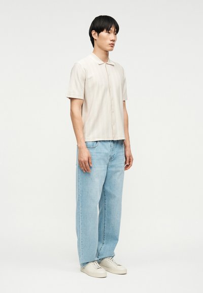 Νεαρός άνδρας στέκεται φορώντας κοντομάνικο μπεζ πουκάμισο με κουμπιά, ανοιχτόμπλε τζιν με φαρδιά μπατζάκια και λευκά αθλητικά παπούτσια σε απλό φόντο.