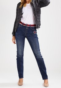 Donkerblauwe spijkerbroek met lichte slijtage, gecombineerd met een zwart gewatteerd jasje, een wit T-shirt en een roodgestreepte riem.