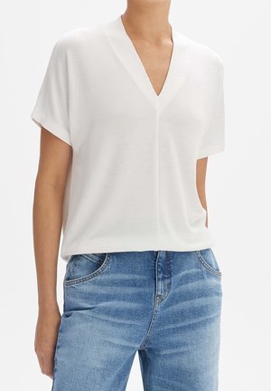 Femme portant un haut blanc à manches courtes et col en V, associé à un jean bleu délavé, debout avec une main dans la poche devant un fond uni.