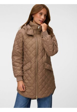 Femme portant une longue veste matelassée marron clair avec poches avant et fermeture éclair, associée à un jean bleu, sur un fond uni.