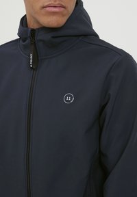 Sötétkék softshell dzseki cipzárral, kapucnival és fehér kör alakú logóval. Simább textúrával és testre szabott fazonnal rendelkezik, letisztult, minimalista stílusban.
