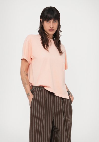 Ανοιχτό ροζ κοντομάνικο μπλουζάκι με άνετη εφαρμογή και στρογγυλή λαιμόκοψη, συνδυασμένο με καφέ παντελόνι με κάθετες ρίγες και πλαϊνές τσέπες.