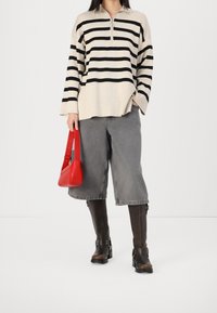 Suéter beige a rayas con cuello de cremallera, jeans grises de pierna ancha y botas de cuero marrón hasta la rodilla. Sosteniendo un pequeño bolso rojo.