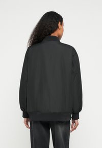 Μαύρο μπουφάν μπομπέρ με λεία υφή, ριμπ μανσέτες και τελείωμα, με ψηλό γιακά και υπερμεγέθη εφαρμογή, εμφανίζεται από πίσω.