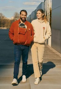 Zwei Personen gehen nebeneinander. Die eine trägt einen strukturierten orangen Fleece mit einer Fronttasche, die andere eine cremefarbene Fleecejacke, beide lässig und gemütlich.