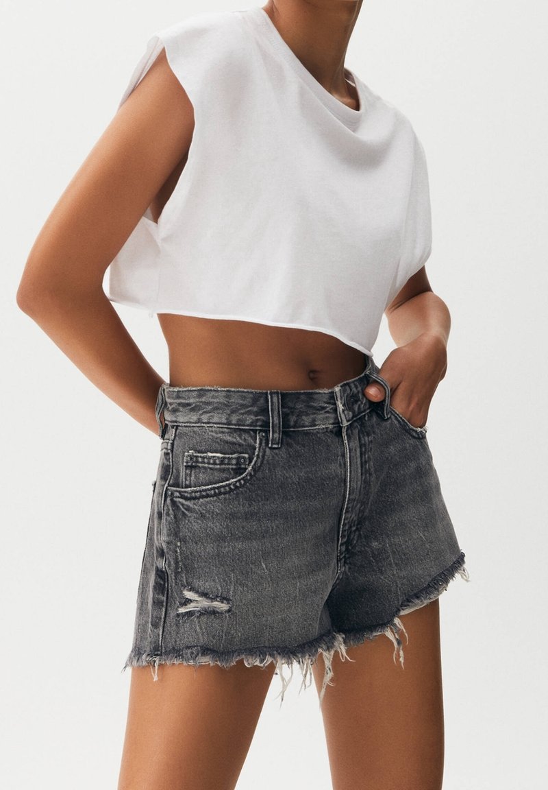 Femme portant un top sans manches blanc court et un short en denim noir déchiré taille haute avec des bords effilochés sur un fond uni.