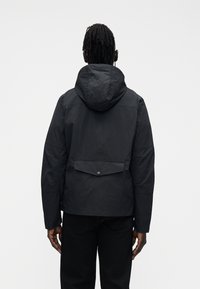 Juoda striukė su gaubtu, pagaminta iš tekstūruotos medžiagos, su nugaros ventiliacijos detalėmis ir spaustukų užsegimu. Sukurta laisvam prigludimui su kasdienio stiliaus estetika.