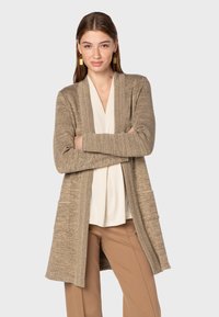 Beige cardigan med öppen front, strukturerad stickad känsla, två framfickor, bärs över en krämfärgad topp. Modellen har armarna i kors och står framför en neutral bakgrund.