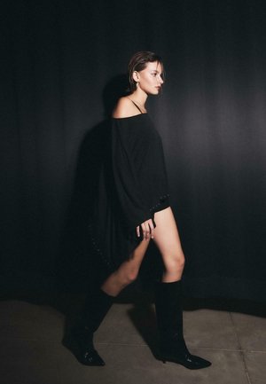 Crna široka majica s otvorenim ramenima, teksturiranim tkanjem i ukrasnim perlicama na rubu. Uparena s crnim čizmama koje sežu do bedara.