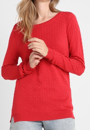 Femme portant un pull rouge texturé à manches longues avec des poignets et un ourlet côtelés, associé à un jean bleu clair sur un fond uni.