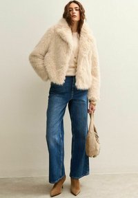 Kvinna som bär en krämfärgad fuskpälsjacka, beige tröja, blå vida jeans, bruna ankelstövlar och håller en beige mockaväskan.