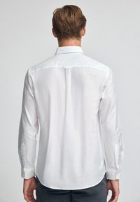 Camicia bianca a maniche lunghe con una trama di tessuto uniforme, dettaglio sul retro, e polsini con bottoni, caratterizzata da delicati accenti di cucitura.