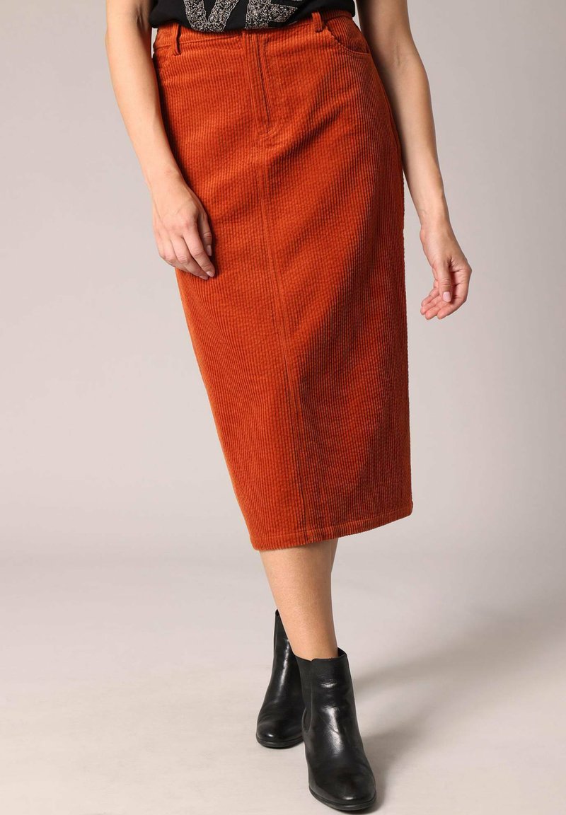 Jupe midi en velours côtelé orange rouillé avec des côtes verticales, dotée d'une taille ajustée et de deux poches latérales, associée à des bottines noires.