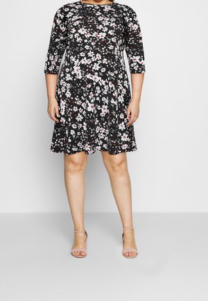 Femme portant une robe noire longueur genou avec des motifs floraux blancs et rouges et des manches trois-quarts, associée à des sandales nude à bride cheville.