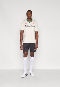 Vyriškas futbolo aprangos komplektas, kuriame yra kreminės spalvos marškinėliai su žaliais ir raudonais akcentais, juodos spalvos šortai, balti aukšti iki kelių kojinės ir balti futbolo bateliai.