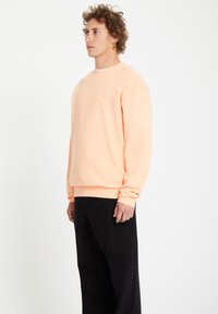 Sweatshirt pêche en mélange de coton, avec col rond et poignets côtelés, associé à un pantalon large noir. Détail de logo simple sur la poitrine.