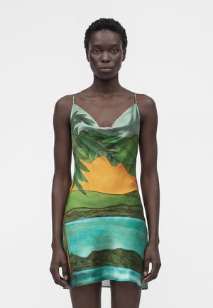 Femme portant une robe sans manches avec un imprimé paysage tropical comportant un feuillage vert, un soleil jaune et de l'eau bleue sur un fond clair.