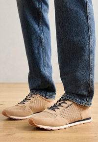 Baskets de sport beige en suede perforé, avec des lacets noirs et des semelles blanches. Associées à un jean en denim bleu.