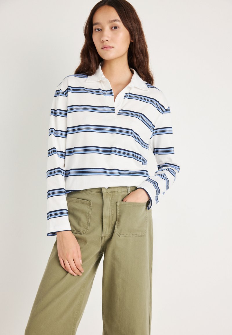 Λευκό πόλο μπλουζάκι με μακριά μανίκια και μπλε οριζόντιες ρίγες, με γιακά. Συνδυάζεται με φαρδιά παντελόνια χακί με μπροστινές τσέπες.