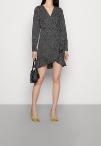 Svart prickig wrapklänning med långa ärmar, volang nederkant och v-ringning. Kombinerad med en liten svart väska och gröna sandaler med remmar.