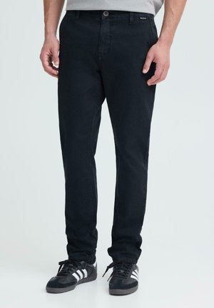 Homme portant un pantalon noir slim et des baskets noires avec des bandes blanches, debout devant un fond clair uni.