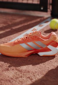 Πορτοκαλί παπούτσι τένις Adidas με γκρι ρίγες, τοποθετημένο σε γήπεδο με χώμα κοντά σε μπάλα τένις και μέρος της γραμμής βάσης.