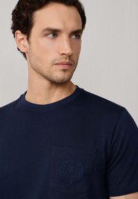 Camiseta de algodón azul marino con cuello redondo y un bolsillo en el pecho con un logo en relieve y diseño de laurel. Textura suave.