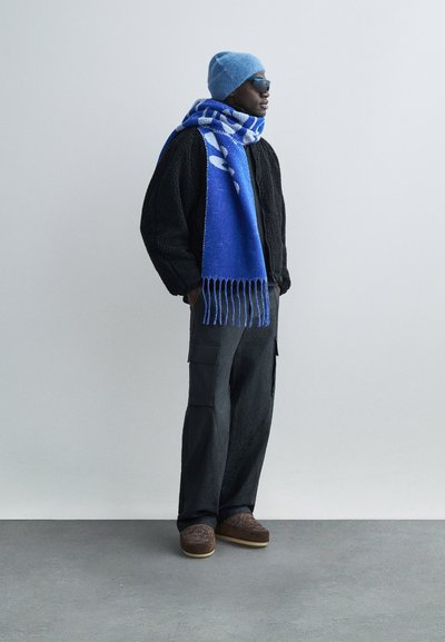 Μαύρο πουλόβερ με υφή, μπλε πλεκτό κασκόλ με κρόσσια, μπλε πλεκτό καπέλο, παντελόνι cargo, καφέ παπούτσια με σχέδια και ανοιχτή σόλα.