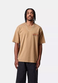 Camiseta de algodón color beige, de manga corta, con un corte relajado, que presenta un pequeño logo impreso en rojo oscuro en el lado izquierdo del pecho. Textura suave.