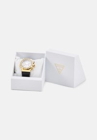 Goldfarbene Chronographuhr mit weißem Zifferblatt und schwarzem Gummiband, präsentiert in einer weißen Geschenkbox mit Guess-Logo, ausgekleidet mit weichem Stoff.