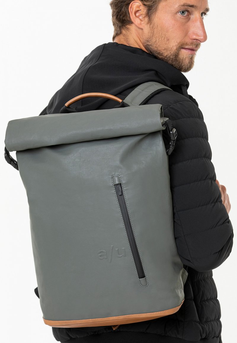 Grau Rolltop-Rucksack mit braunen Lederakzenten und einem seitlichen Reißverschluss, aus glattem Material gefertigt und mit verstellbaren Trägern.