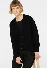 Schwarze Strickjacke mit V-Ausschnitt, vorderer Knopfleiste und gerippten Bündchen. Präsentiert mit einer glatten Textur und lockerer Passform, kombiniert mit einem dunklen Oberteil.