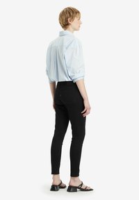 Camisa de rayas azul claro con un corte relajado, jeans ajustados negros y sandalias de tacón negras. Mangas enrolladas visibles y un sutil detalle en la espalda.
