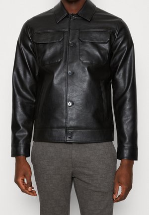 Mand iført sort læderjakke med knapper og brystlommer samt grå mønstrede bukser, stående med armene afslappet ned langs siderne.