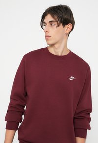 Burgundský Nike mikina s dlhými rukávmi, rebrovanými manžetami a okrúhlym výstrihom. Na ľavej strane hrude sa nachádza malé biele logo.