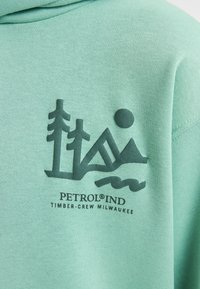 Felpa verde turchese con una grafica verde scuro di alberi, montagne e un sole, con il testo "PETROL IND" sottostante.