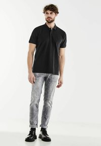Black polo shirt with short sleeves, paired with light gray faded jeans and black lace-up boots. Model stands against a white background.
