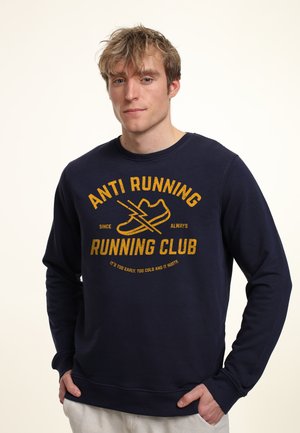 Mørkeblå sweatshirts lavet af et blødt stof, med et gult trykt grafik, der lyder "ANTI RUNNING CLUB" med et skodesign og slogan.