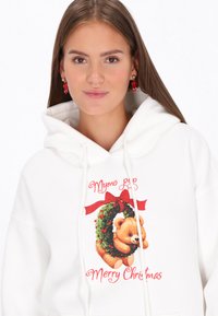 Fehér kapucnis pulóver, ünnepi macimintával, mely koszorút és piros szalagot ábrázol. A nyomat piros "Boldog Karácsonyt" feliratot tartalmaz.