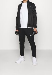Chaqueta deportiva negra con rayas blancas, camiseta blanca, pantalones negros con el logo de Nike y zapatillas blancas, sosteniendo mancuernas.