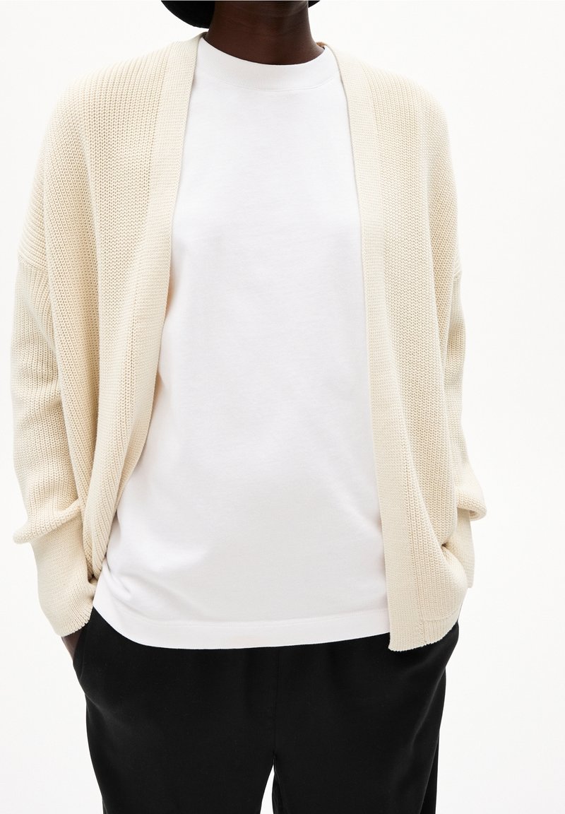 Béžový pletený kardigan s otevřeným předním dílem, nošený přes bílou tričko s kulatým výstřihem. Má volné rukávy a uvolněný střih. Stylizováno s černými kalhotami.