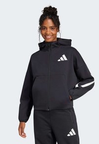 Felpa con zip nera realizzata in tessuto morbido, dotata di cappuccio, logo bianco e dettagli a righe sulle maniche; vestibilità comoda con tasche frontali.