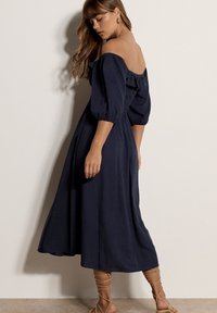 Femme aux longs cheveux portant une robe midi bleu marine épaules dénudées avec manches bouffantes et des sandales beige à lacets, vue de profil sur un fond uni.