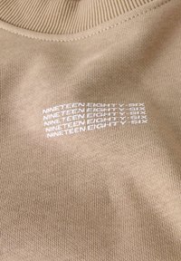 Beige strikket stof med hvid tekst gentaget i fem rækker: "NINETEEN EIGHTY SIX" på et stykke tøj.